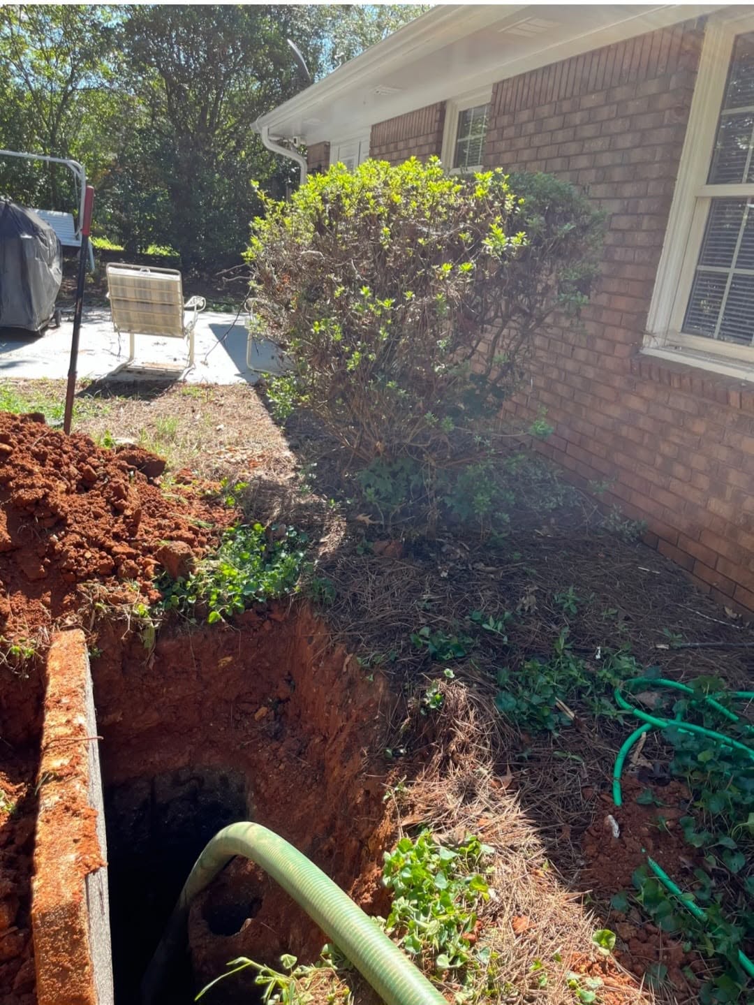 Excavated backyard pit with a pump hose beside a brick home