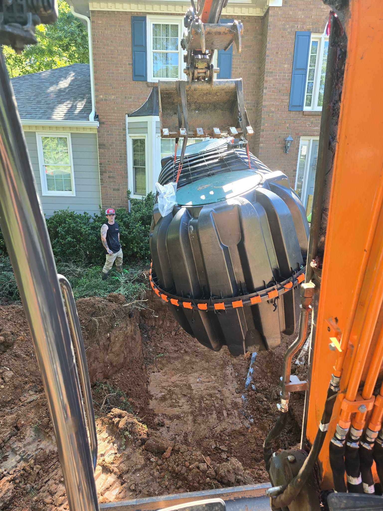 Excavator lowering a black septic tank with green lids into a front-yard pit